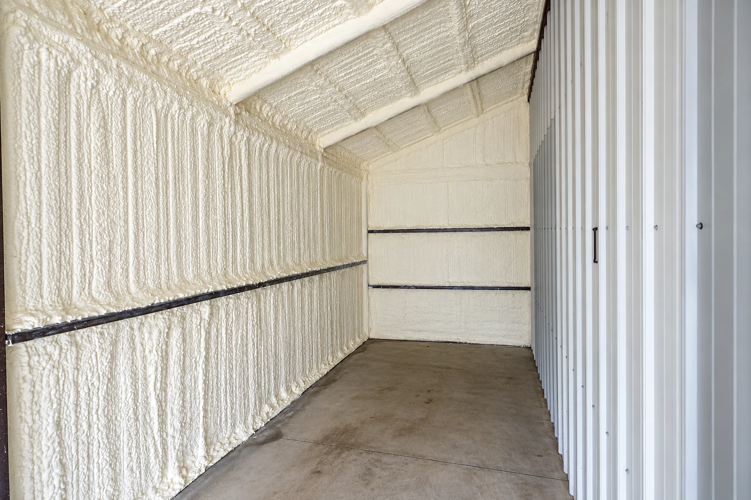 A narrow room has white spray foam covering the walls and sloped ceiling beside a corrugated metal wall.