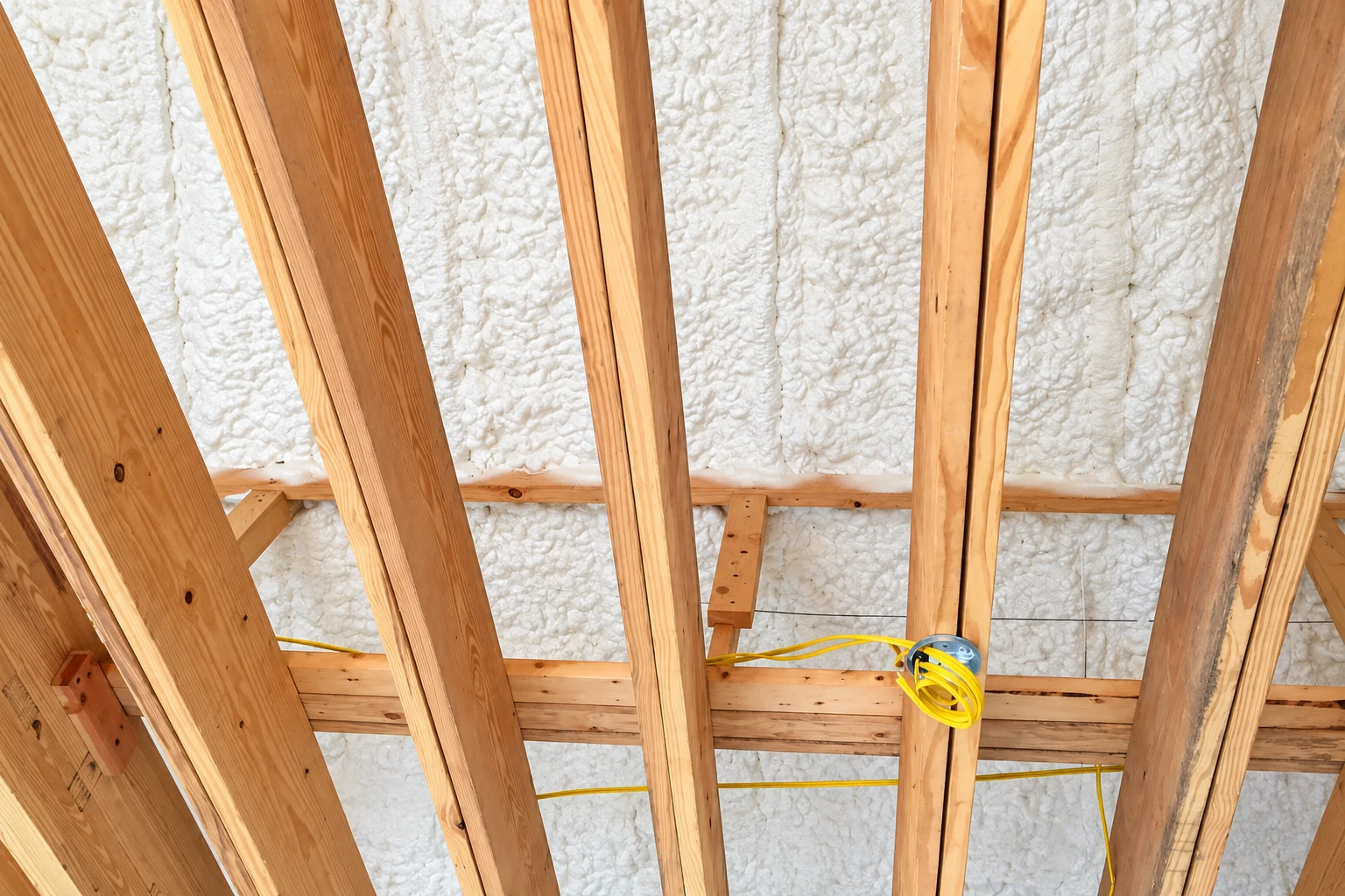White spray foam fills the roof cavities between exposed wooden rafters, with yellow electrical wiring visible.