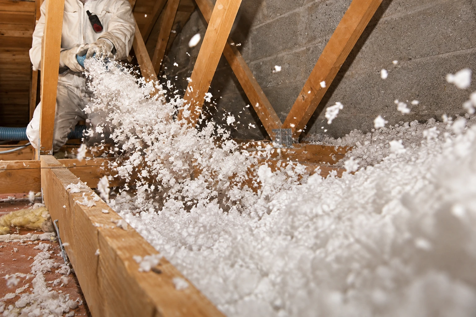 A worker sprays loose white insulation through a hose into the open bays of a wooden attic.