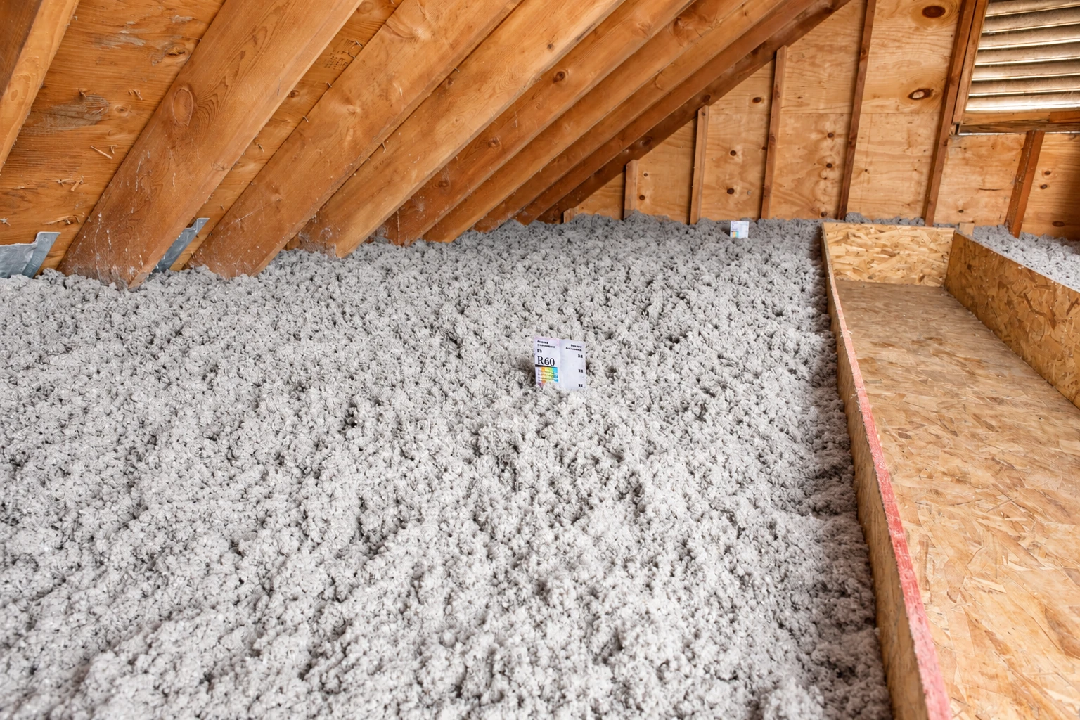 A wooden attic floor is covered with a deep layer of gray blown-in insulation beside a narrow walkway.