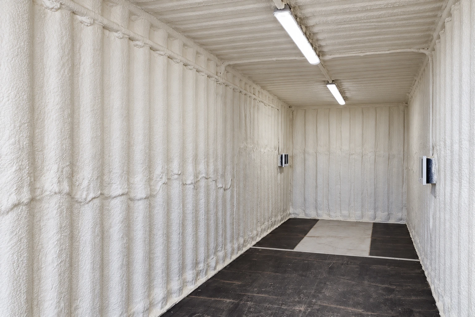 A long commercial interior is coated with white spray foam on its walls and ceiling, with two overhead lights.