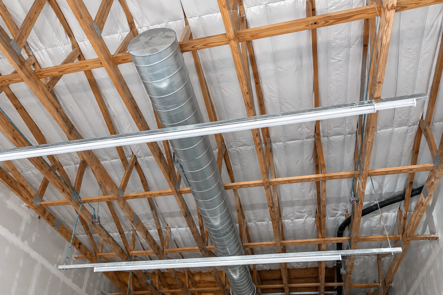 Exposed wooden roof trusses filled with white insulation, with metal ductwork and long suspended light fixtures.