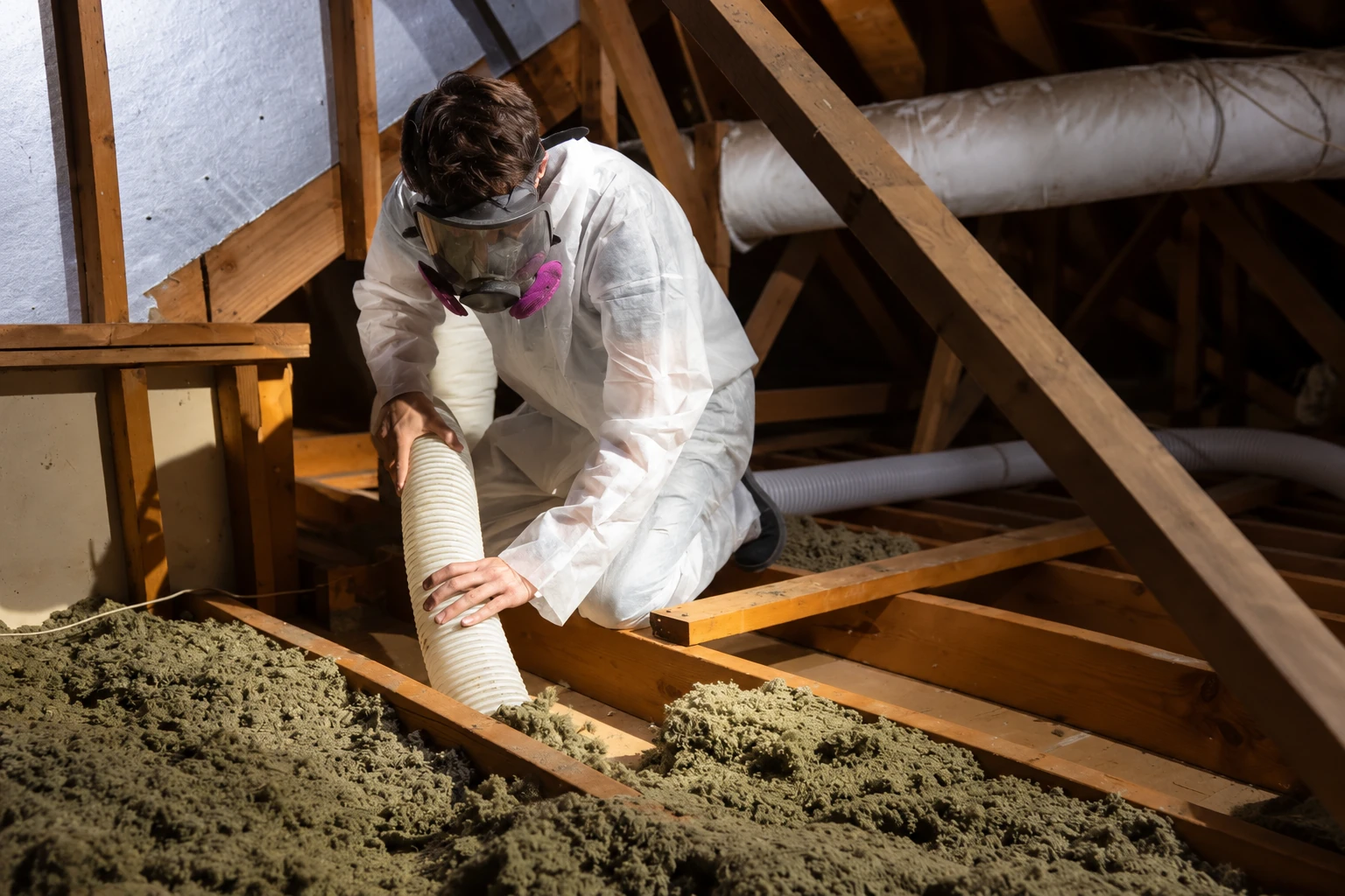 Worker in protective coveralls and a respirator removes loose attic insulation using a flexible vacuum hose.